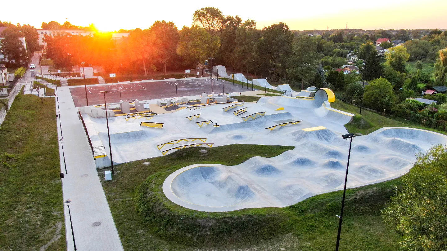Terenu skatepark z licznymi rampami, przeszkodami i betonowymi muldami oraz w tle boisko do koszykówki.
