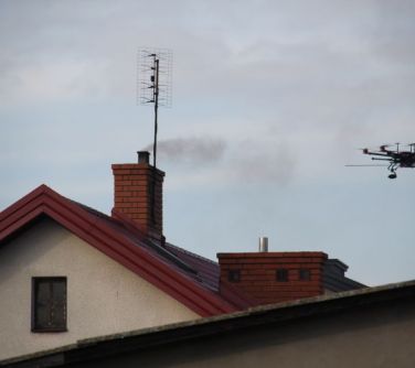 Dron na tle nieba. Po lewej komin budynku. Z kominu leci dym