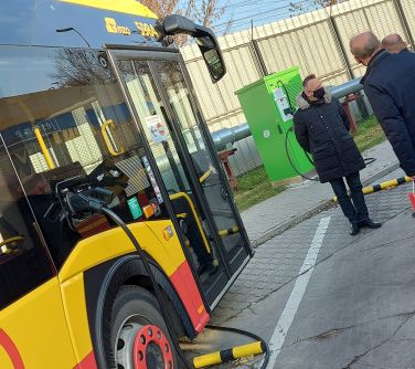 Autobus elektryczny podłączony do ładowania. Delegacja z urzędu obserwuje.