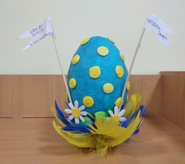 Niebieskie jajo w żólte kropki, wykonane z plasteliny. U podstawy kwiatki i pióra.