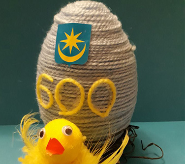 Jajko oplecione sznurkiem z naklejonym herbem miasta i cyfrą 600, przed nim kurczak.