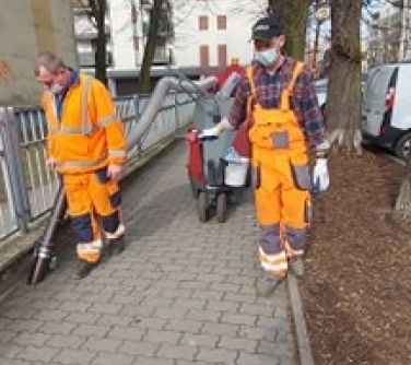 Dwóch pracowników ZGK w pomarańczowych uniformach. Sprzatają chodnik przy pomocy nowego urządzenia.