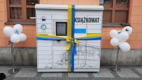 zdjęcie książkomatu stojącego na chodniku przed MBP w MIńsku Mazowieckim, opleciony niebiesko-żółtą wstęgą, po obu bokach...