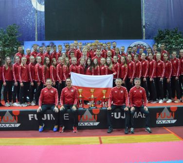 Oświetlone wnętrze. Duża grupa zawodników w czerwonych bluzach. Po środku trzymają flage bialo-czerwoną