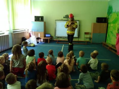 Wnętrze przedszkola, sala. Dzieci siedzą, przed nimi strażak