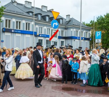 Dzień, pochmurno. Na placu zgromadzone osoby w przebraniach z poprzedniej epoki. Na przodzie mężczyzna we fraku i kapeluszu