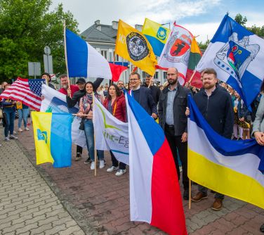 Dzień, pochmurno. Duża grupa osób. Trzymaja flagi miast partenrskich