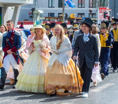 Dzień, słońce. Po ulicy maszeruja osoby w przebraniach. Na przodzie dwie kobiety w żółtych długich falbaniastych sukienkach