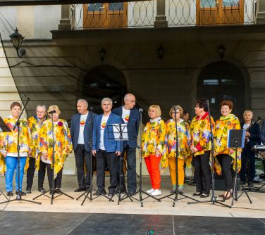 Na scenie osoby. Po środku mężczyźni. Po obu stronach seniorki w żółtych w kwiaty bluzkach