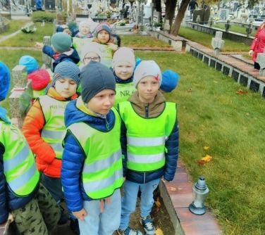 grupa dzieci w odblaskowych kamizelkach odwiedza cmentarz, na pierwszym planie dwóch chłopców