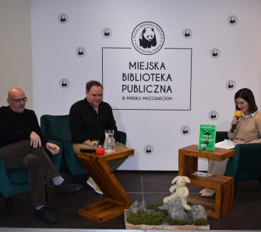 Dwóch mężczyzn i kobieta siedzą  na fotelach przy stoliku. W tle baner Miejskiej Biblioteki Publicznej