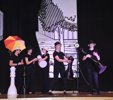 dziewczyny stoją na scenie, wielkie pionki do szachów, kolorowy parasol w tle