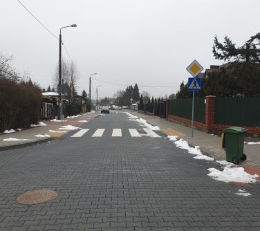 Na zewnątrz, pochmurny dzień. Skrzyżowanie, po lewej na chodniku zielony pojemnik na śmieci, w tle po lewej stoi samochód