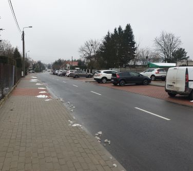 ulica, zdjęcie zrobione na chodniku, po lewej stronie siatka posiadłości, po prawej stronie parking na którym stoja samochody