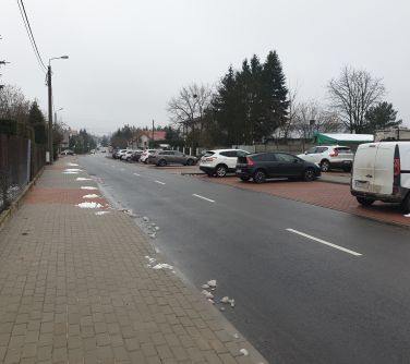 Na zewnątrz, pochmurny dzień. Ulica, zdjęcie zrobione na chodniku, po lewej siatka, po prawej parking z samochodami