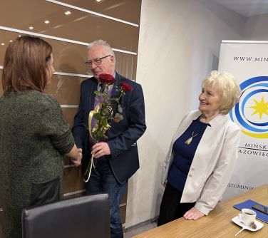 mężczyzna gratuluje kobiecie, trzyma trzy czerwone róże trzyma w ręku, obok stoi kobieta