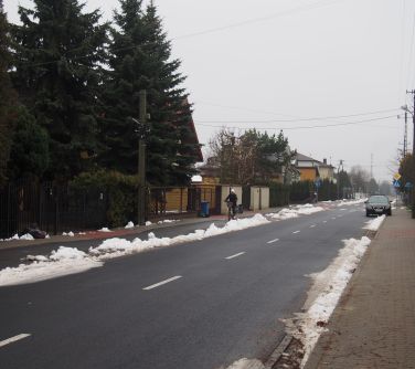 ulica, snieg po obu stronach krawęzników, po prawej stronie stoi samochód, po lewej stronie osoba jadąca rowerem po chodniku