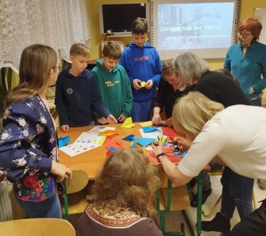 na około stoły na którym leży kolorowe origami, stoją dzieci, kobiety, kobieta tyłem nachyla się i robi zdjęcie