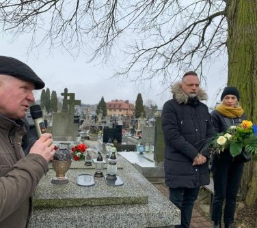 mężczyzna po lewej stronie przemawia do mikrofonu, po prawej stronie kobieta stoi z mężczyzną, kobieta trzyma kwiaty