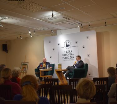 na scenie dwóch mężczyzn siedzi w fotlach, jeden przemawia, na zdjęciu od tylu publicznośc siedząca na krzesłach