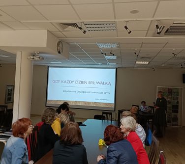 osoby siedzą przy stole przed nimi na ekranie wyświetlane slajdy