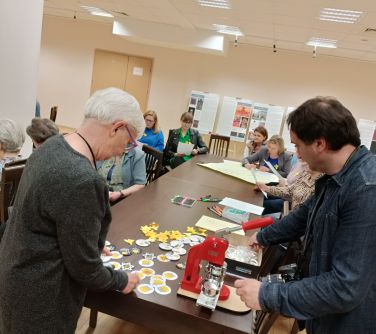 po lewej kobieta nachyla się nad stołem po prawej mężczyzna prygotowujący sie do zrobienia przypinek, w tle osoby siedzące...