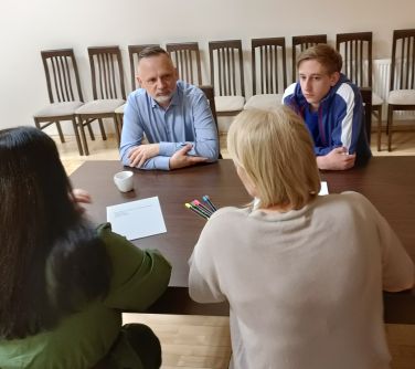 cztery osoby siedzą przy stole rozmawiają, na stole leżą kartki i kolorowe mazaki, w tle stoja krzesła pod ścianą