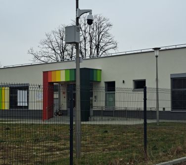 zdjęcie latarni przy ogrodzeniu z zamontowanym monitoringiem, w tle przedszkole