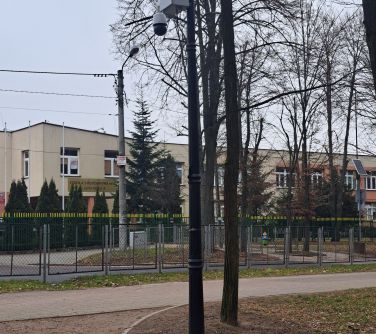 zdjęcie alejki przy której stoi latarnia  z zamontowanym monitoringiem, w tle ogrodzenie i szkoła
