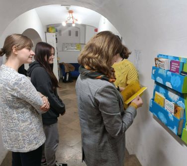 cztery kobiety stoją przy ścianie z zaczepionymi przegródkami na ulotki, jedna z nich przegląda ulotkę