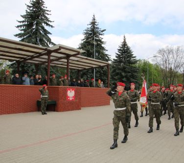 żołnierze maszerują w defiladzie, po lewej stronie osoby na murowanym pdwyższeniu, na murze z przodu godło Polski