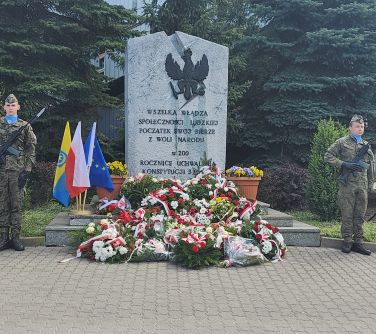 pomnik konstytucji 3 maja, przed pomnikiem kwiaty, po lewej stronie flagi, po obu stronach stoja żolnierze