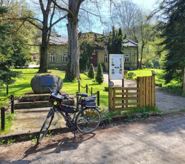 zdjęcie stojącego roweru przed pomnikiem z kamienia, w tle drzewa i domek