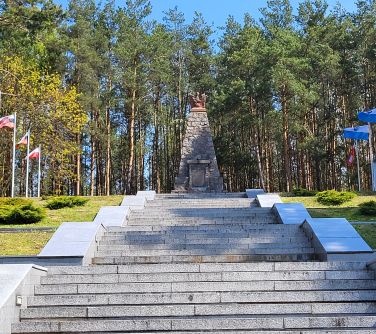 zdjęcie schodów prowadzących do pomnika w kształcie stożka, po obu stronach flagi, w tle las