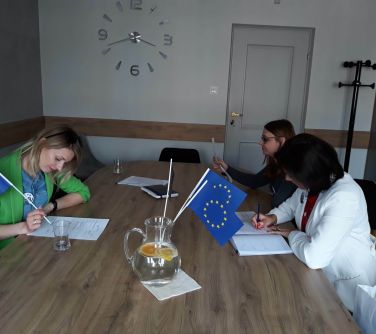 kobiety siedzą przy stole, czytają, na stole flagi UE, w tle zegar wisi na ścianie