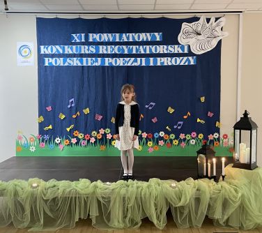 dziewczynka stoi na scenie, po prawej stronie dwa lampiony, w tle granatowa płachta z tytułem głównym XI Konkurs...