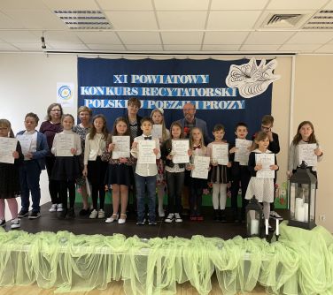 osoby stoją na scenie, po prawej stronie dwa lampiony, w tle granatowa płachta z tytułem głównym XI Konkurs Recytatorski...