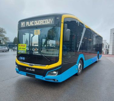 elektryczny autobus stoi na ulicy w tle filar budynku po prawej, po lewej stoja samochody na parkingu