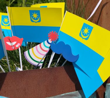 papierowe flagi Mińska Mazowieckiego wbite w donicę z ziemią przed nimi papierowa czapeczka urodzinowa i papierowe usta