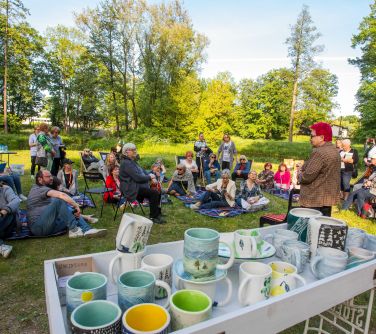 piknik na trawie, osoby siedzą na leżakach, kocach, na przodzie stolik z kubkami i dzbankami