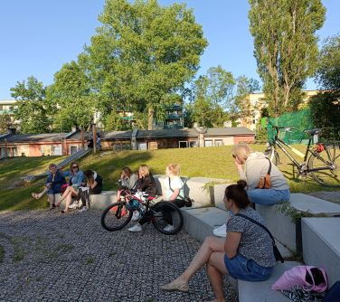 osoby siedzą na betonowych dużych schodach w półkolu, przed nimi rower