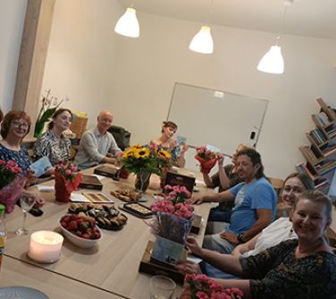 grupa osób siedzi przy stole na którym stoją kwiaty, ciastka, owoce