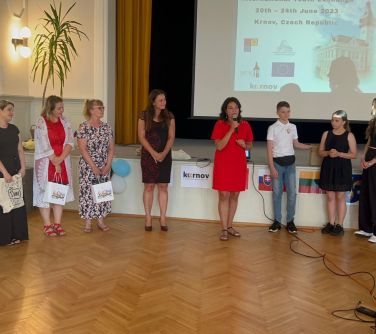 grupa osób w sali ze sceną, w środku kobieta w czerwonej sukience mówi do mikrofonu, po prawej stoją kobiety z torbami...
