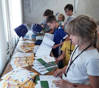 dzieci stoją przy blacie na którym przeglądają rozłozone ksiązki i czasopisma, po środku w szklance małe flagi unii...
