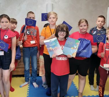 grupa dzieci pozuje do zdjęcia trzymając w dłoniach małe flagi ue i worki, uśmiechnięty chłopiec z przodu na środku trzyma...