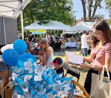 po prawej kobieta przegląda publikację, przed nią na stole balony, wiatraczki, koszyk z gadżetami po lewej dzieci