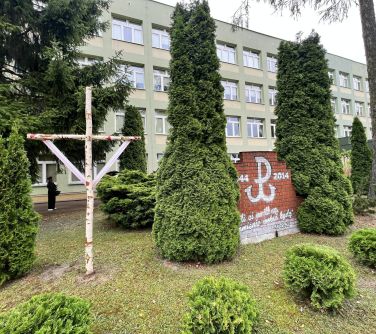 ogórd przed szkołą, krzyż i ściana z symbolem powstańczym