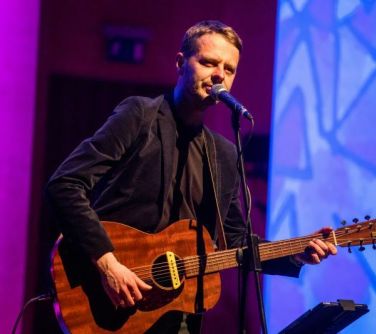 mężczyzna stoi na scenie przed mikrofonem, gra na gitarze