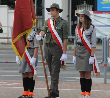 Trójka młodych harcerzy stoi w poczcie sztandarowym, chłopiec w środku trzyma sztandar