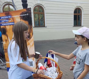 dwie dziewczynki stoją przy stole, wyjmują z koszyka lizaki i flagę, w tle baner UE